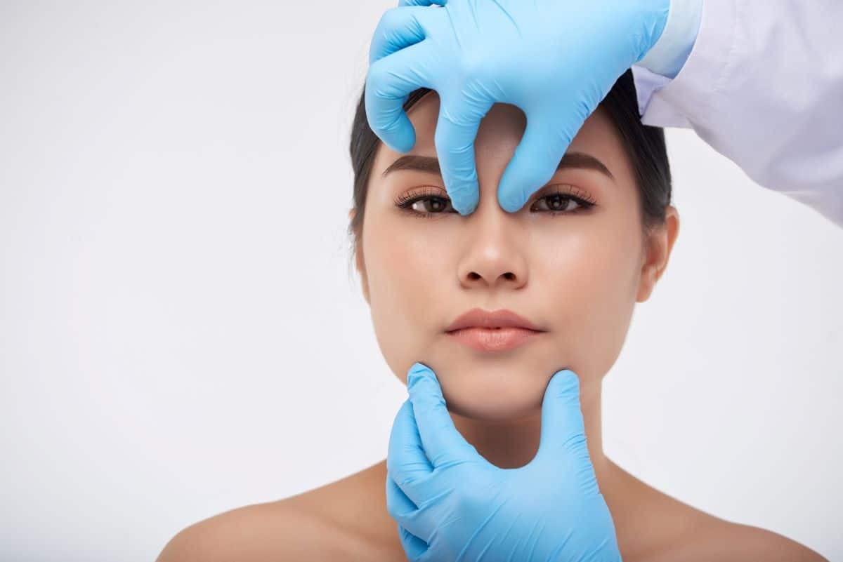 gloved hands of an ENT gently pinch the bridge of the nose of a female patient who is looking directly at the viewer of the image