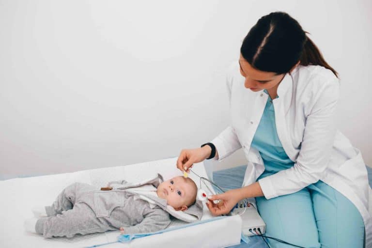 A baby in a onesie lying on a bed while a doctor places electrodes on it's head.